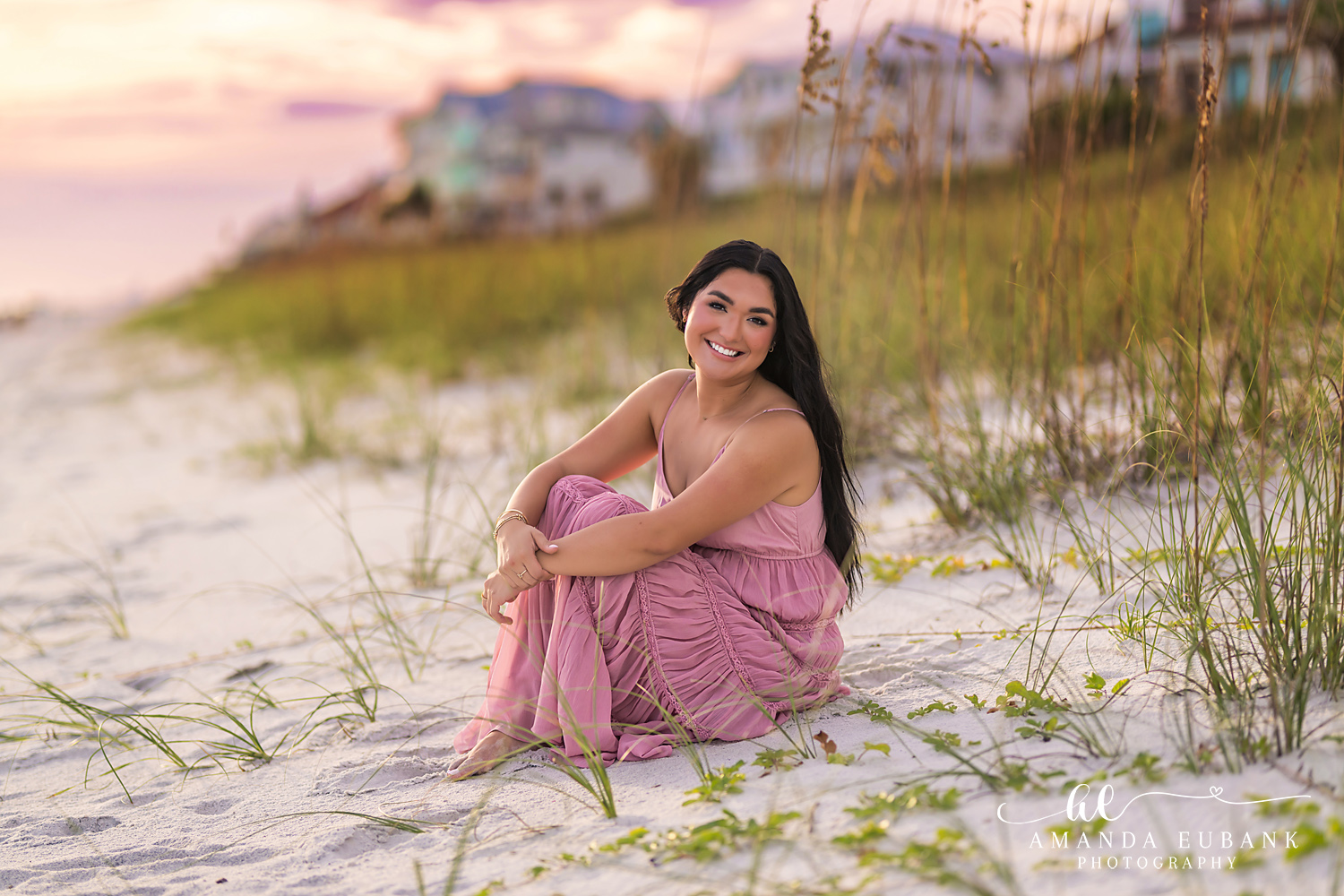 Rosemary Beach Senior Photos | Sitting in the sea oats Rosemary Beach senior photographer: senior girl in a pink maxi dress sitting in the sea oats on a sand dune with beach houses behind her