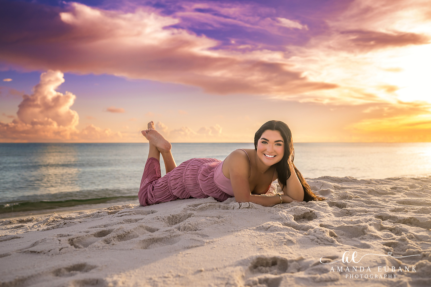 Rosemary Beach Senior Photos | Lying on the sand under a colorful sky Rosemary Beach senior photographer: senior girl in a pink dress lying on her stomach on the sand with feet raised under a colorful sky