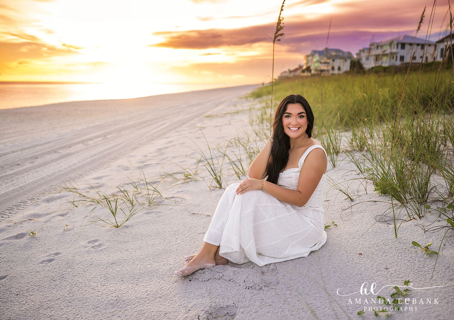 Rosemary Beach Senior Photos | White dress in the sea oats Rosemary Beach senior photographer: senior girl in a white dress sitting in the sea oats on the dunes with beach houses and a bright sky behind