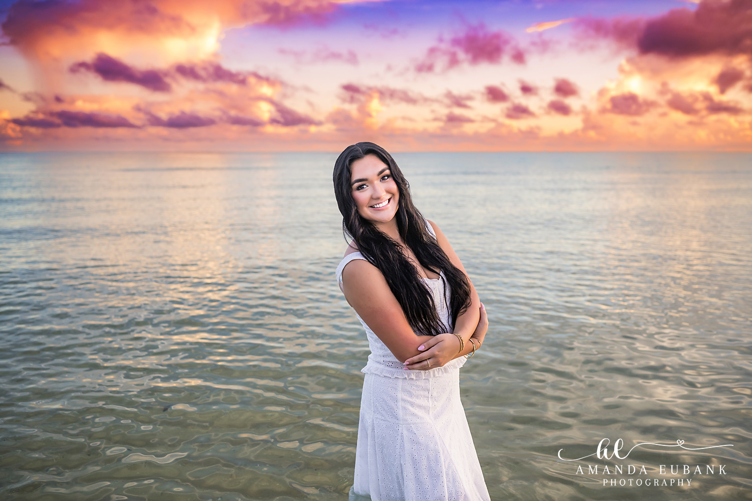 Rosemary Beach senior photographer: senior girl in a white dress standing in the Gulf with arms crossed and a big smile under a colorful sky